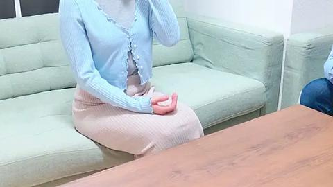 Media: Video of a woman in a light blue cardigan, pink skirt, and gray top seated on a light green sofa, holding a pink phone, with a wooden table in the foreground.