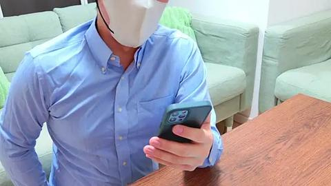 Media: Video of a person wearing a light blue button-up shirt, white mask, and holding a smartphone, sitting on a wooden table in a living room with green couches.