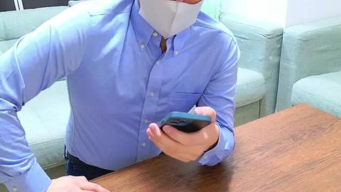 Media: Video of a person wearing a light blue button-up shirt, face mask, and gloves, holding a black smartphone over a wooden table. Background includes a light green sofa and white walls.