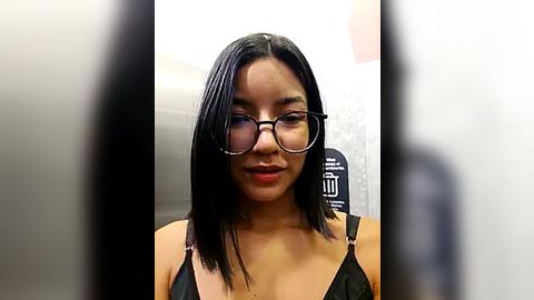 Media: Video of a young Asian woman with straight black hair, wearing glasses and a black top, smiling slightly. Background shows blurred subway car seats.