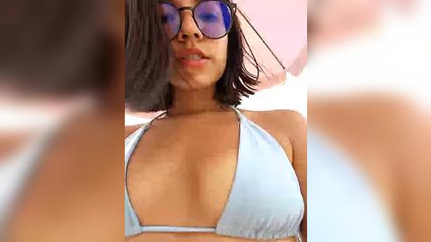 Media: Video of a woman with medium brown skin, wearing a light blue bikini top, short black hair, and round sunglasses. She stands under a pink umbrella, with blurred figures in the background.