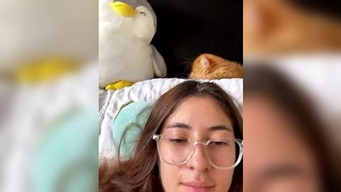 Media: Video of a young woman with fair skin, brown hair, and glasses, lying on a bed with a penguin plushie, an orange cat, and a white blanket.