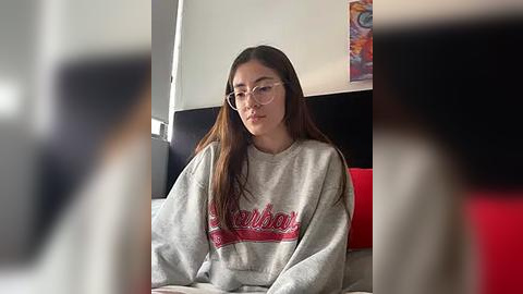 Media: Video of a young woman with long dark hair, wearing glasses, a grey sweatshirt with red letters, sitting on a bed in a bright room.