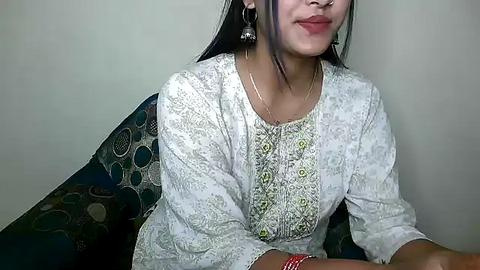 Media: Video of a young woman with light brown skin and straight black hair, wearing a white embroidered kurta, seated on a patterned sofa, smiling.
