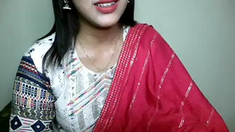 Media: Video of a woman with medium skin tone, straight black hair, and wearing a colorful, patterned white blouse with geometric designs, draped in a vibrant red saree. She smiles, with a subtle, delicate gold necklace.