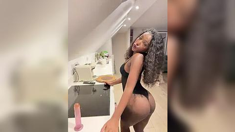 Media: Video of a slender, dark-skinned woman with curly hair in a tight black bodysuit, posing provocatively in a modern, minimalist kitchen. She holds a tray with a pink dildo and a bowl.