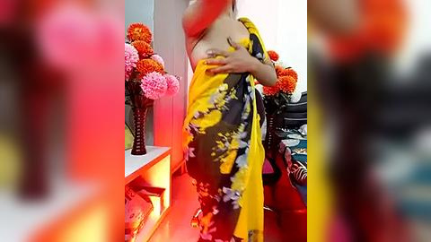 Media: Video of a woman in a yellow and black floral sari, holding her breasts, in a brightly lit room with pink and orange flowers, blurred in the foreground.