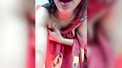 Media: Video of a woman with medium brown skin, wearing a maroon sari with yellow and green patterns, revealing her bare chest, her mouth open in a loud expression, red lighting, blurred background.