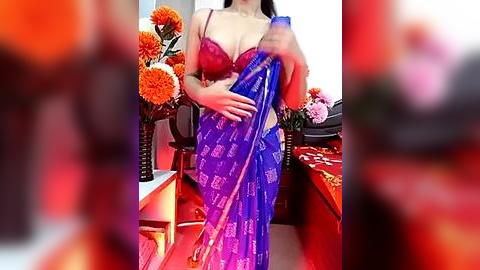 Media: Video of a woman in a red lace bra and purple sari, holding a blue sari. Background features orange flowers, red table, and blurry figures.