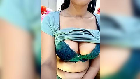 Media: Video of a South Asian woman with medium brown skin, wearing a teal lace bra, lifting her shirt to reveal her large breasts. Background includes blurred floral arrangements.