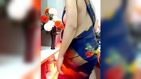 Media: Video of a woman with fair skin wearing a deep blue, floral-patterned, sleeveless dress with a plunging back. She stands in a brightly lit room with orange and white flowers in a vase on a white counter.