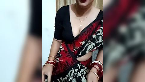 Media: A blurred video of a woman in a black and red floral sari with a plunging neckline, gold bangles, and a gold necklace, indoors with beige curtains in the background.