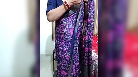 Media: A video of a woman in a vibrant purple saree with intricate floral patterns, wearing a blue blouse and multiple red bangles, standing in a room with a red couch and wooden furniture in the background.