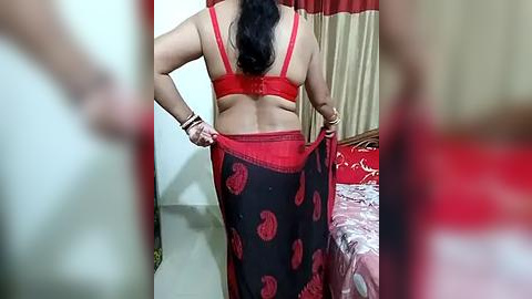 Media: A video shows a woman with dark hair and a red bra, adjusting a black sari with red floral patterns in a bedroom with beige curtains, red bedspread, and a brown headboard.