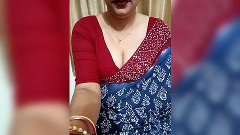 Media: Video of a woman with medium skin tone wearing a red blouse and blue sari with white patterns, red lipstick, and gold jewelry, in a red and gold room.