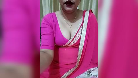 Media: A video shows a woman in a vibrant pink saree with a plunging neckline, exposing ample cleavage, and wearing a gold necklace. The background features beige curtains.