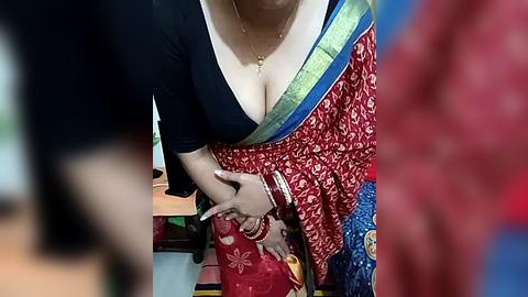 Media: A close-up video of a woman in a deep neckline blouse and red saree with intricate patterns, showing cleavage, wearing gold bangles and a gold necklace, in a cluttered room.