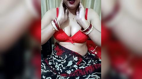 Media: Video of a light-skinned woman with large breasts, wearing a red lace bra, multiple bangles, and a colorful saree, her hands touching her neck. Background is blurred, featuring a beige curtain.