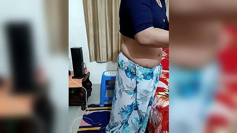 Media: Video of a person with a round belly, wearing a blue shirt and floral-patterned pajama pants, standing in a cluttered room with a TV, fan, and a red bedspread.