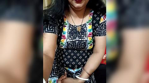 Media: Video of a woman with medium skin tone, wearing a black and white patterned dress with colorful geometric designs, black hair, smiling, in a blurred background.