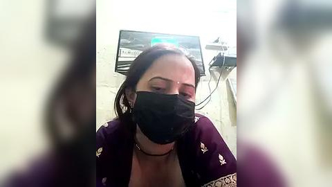 Media: A video of a woman with short brown hair wearing a black face mask, a purple robe, and a black choker, sitting in a dimly lit room with medical equipment in the background.