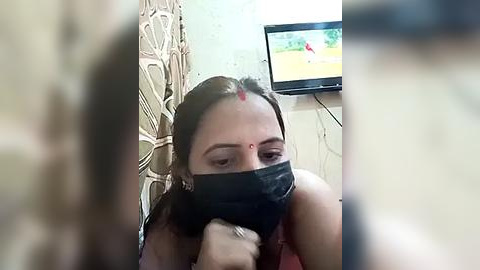 Media: A video of a woman with brown skin and long hair, wearing a black mask and a red bindi, sitting in a dimly lit room with a TV in the background.