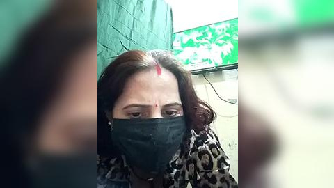 Media: A video shows a woman with dark hair and a red bindi on her forehead, wearing a black mask, standing in front of a green wall. The background includes a green TV screen displaying white text.