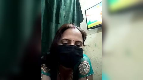 Media: Video of a woman with light brown skin and dark hair, wearing a green saree, black face mask, and gold jewelry, in a dimly lit, simple room with green curtains and a TV showing green text.