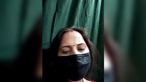 Media: Video of a woman with dark skin, wearing a black mask, red dot on her forehead, and a green saree, blurred background.