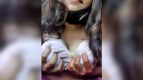 Media: A blurred video of a person with long, wavy brown hair, wearing a white shirt, and black choker, their hands grasping a green object.