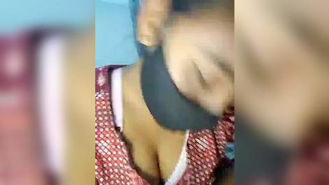 Media: Video of a woman with medium brown skin, wearing a red and white patterned blouse with low-cut neckline, partially obscured by a black mask, face slightly blurred, background indistinct.