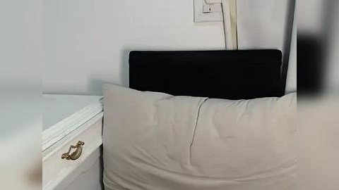 Media: Video of a minimalist bedroom featuring a black upholstered headboard, two white pillows, and a white nightstand with a brass handle. The walls are painted white, and an electrical outlet is visible on the wall.