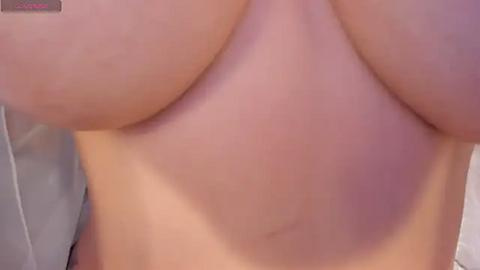 Media: Video of a nude person with light skin, showcasing their ample, natural breasts, with visible areolas and nipples. The background is blurred, featuring a white wall. The image captures a natural, unembellished depiction of the human body.