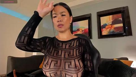 Media: Video of an East Asian woman with medium skin tone, black hair, and a slender physique, wearing a sheer black top with geometric patterns, seated on a black couch, in a modern living room with colorful abstract paintings on the wall.