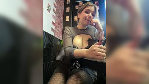 Media: Video of a curvy, light-skinned woman with tattoos, wearing a tight, grey top and fishnet stockings, sitting in a brick-walled room, holding a cup.