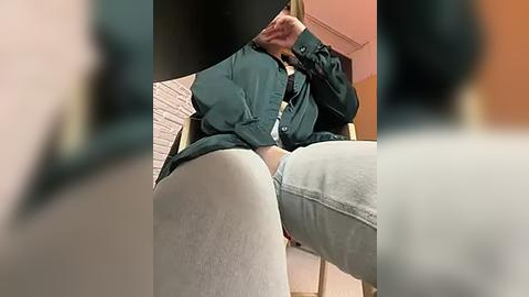 Media: Video of a person in a green jacket, white pants, sitting on a chair with legs spread wide, partially visible genitals. Background shows a blurred, warm-colored room.