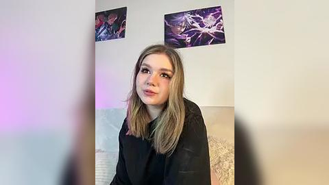 Media: Video of a young woman with straight, shoulder-length blonde hair, wearing a black shirt, standing against a light-colored wall with purple and white abstract art prints above.