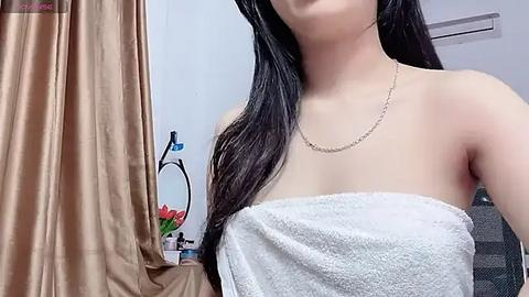 Media: Video of a young Asian woman with long black hair, wrapped in a white towel, standing in a bathroom with beige curtains and a floral arrangement on the left.