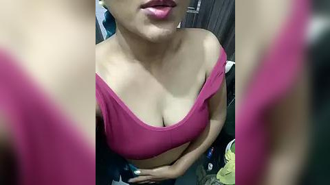 Media: A close-up video of a woman with light brown skin, wearing a low-cut, off-the-shoulder magenta top, showcasing ample cleavage. Her lips are slightly open, and the background shows a blurred room with wooden furniture.