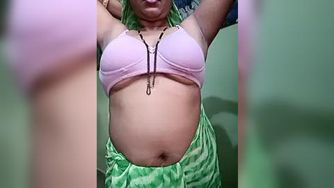 Media: Video of a pregnant woman with light brown skin, wearing a pink bra and green sarong, lifting her arms, showcasing her belly. Background is blurred, focusing on her torso.