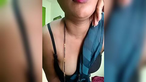 Media: Video of a woman with light brown skin, wearing a black lace bra and a blue blouse, with a silver necklace, standing indoors.