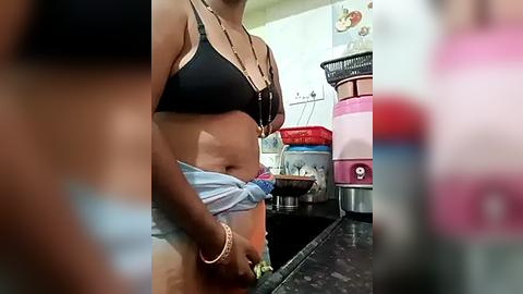 Media: Video of a woman in a kitchen, wearing a black bra and light blue shorts. She is holding up her shorts. Background includes a pink and white blender, a bowl, and a white wall.