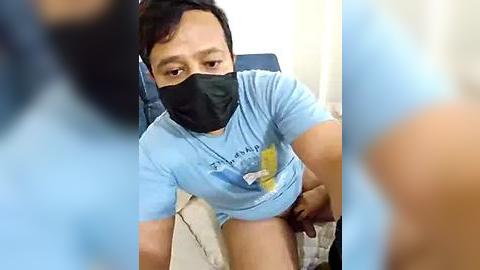 Media: A video of a young Asian man in a light blue t-shirt and black mask, sitting on a bed, possibly in a hospital or clinic setting.