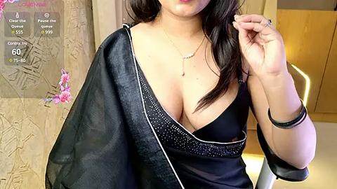 Media: Video of a curvaceous woman with long dark hair, wearing a low-cut black saree with a gold necklace, standing in a modern indoor setting with textured walls and wooden accents.