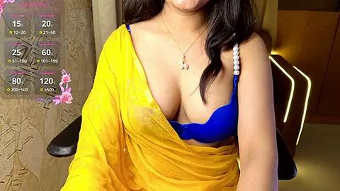 Media: Video of a woman with medium skin tone and long black hair, wearing a yellow sari and a bright blue bra, sitting indoors.