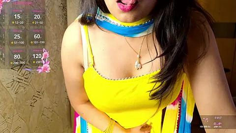 Media: Video of a woman with long black hair, wearing a bright yellow kurta with a blue scarf, pink lipstick, and a gold necklace. Background shows a textured wall and a digital health app interface.