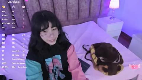 Media: A video of a young woman with long black hair, wearing a colorful graphic sweatshirt, sitting on a bed with a brown dog, in a dimly lit bedroom with purple lighting.