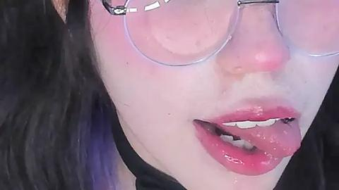 Media: Video of a close-up of a woman's face with fair skin, wearing round glasses, pink lipstick, and a black choker. Her long black hair is visible on the left.