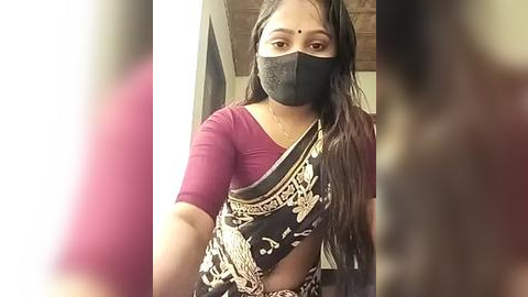 Media: Video of an Indian woman with long black hair, wearing a black face mask, a maroon top, and a black saree with gold floral patterns. Background shows blurred people and a wooden ceiling.