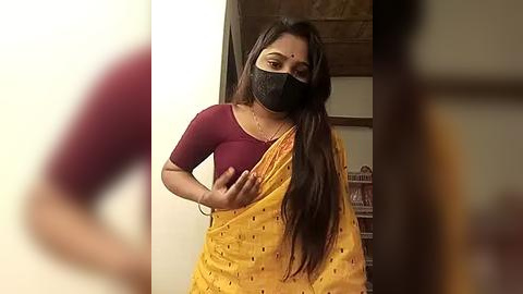 Media: A video shows a South Asian woman with long black hair, wearing a maroon top, yellow sari with subtle patterns, and a black face mask, standing indoors with a blurred background.